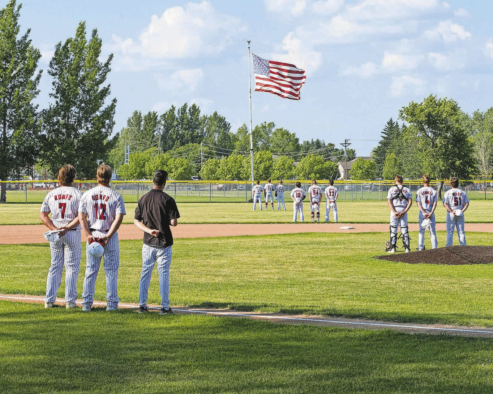 Players stand facing the flag as the National Anthem plays at the start of the game against Midway/Minto on June 30 in Langdon. PHOTO BY LARRY STOKKE.