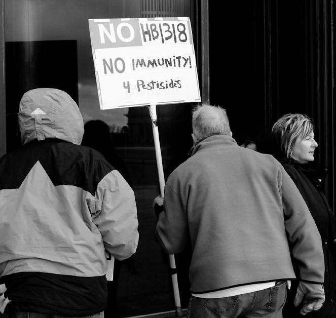 Protesters opposing a bill that would raise the standard for proving liability for lawsuits involving pesticides peer through the windows of the Capitol on April 14, 2025. (Michael Achterling/North Dakota Monitor)