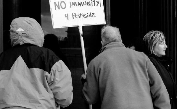 Protesters opposing a bill that would raise the standard for proving liability for lawsuits involving pesticides peer through the windows of the Capitol on April 14, 2025. (Michael Achterling/North Dakota Monitor)